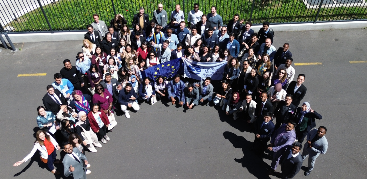 Daudz cilvēku sastājušies puslokā, rokās Eiropas Savienības un Erasmus Mundus Asociācijas karogs