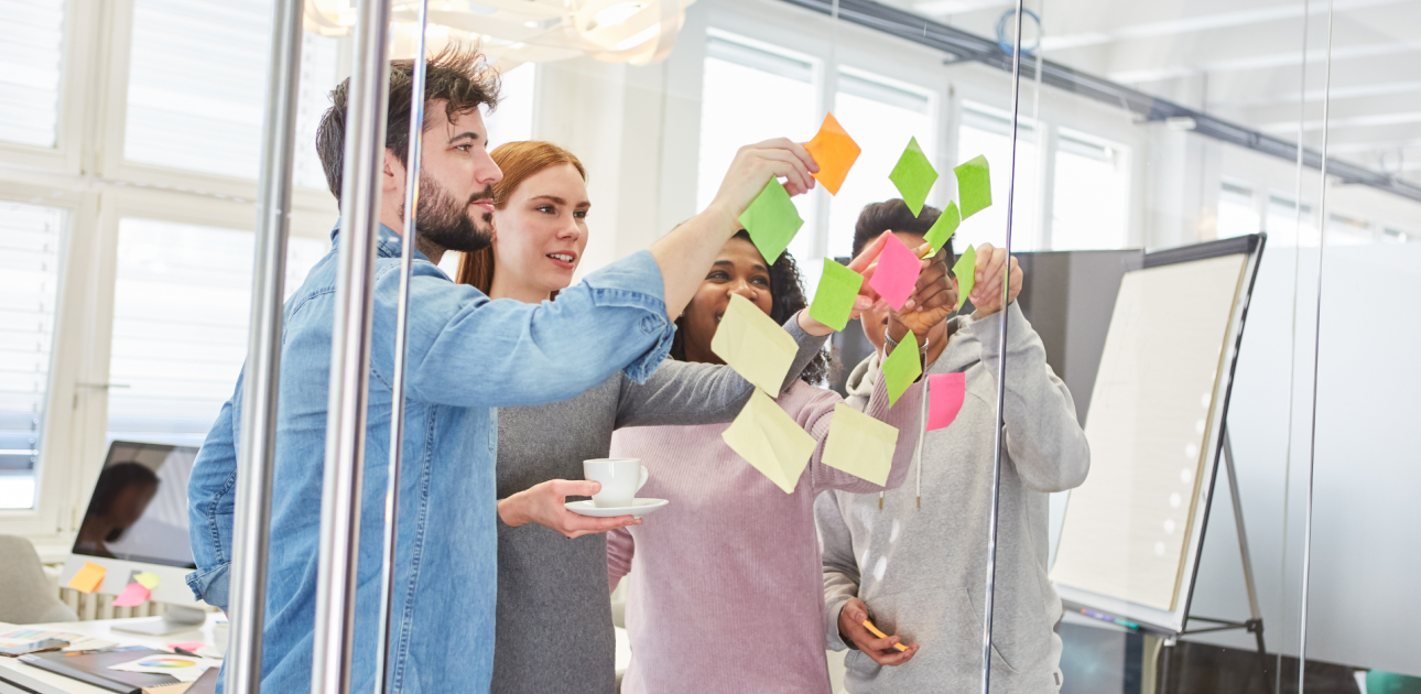 četri cilvēki stāv pie stikla sienas, līmē krāsainas līmlapiņas