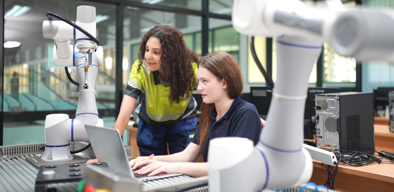 jauna meitene sēž pie datora, viņai blakus kaut ko stāsta pedagogs sieviete, uz galda blakus stāv robototehnika, viņi atrodas mācību darbnīcā