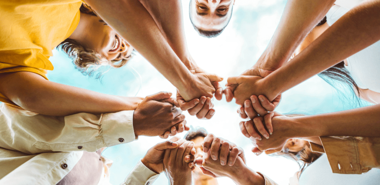 Cilvēku grupa aplī, turot kopā saliktas rokas pret debesīm