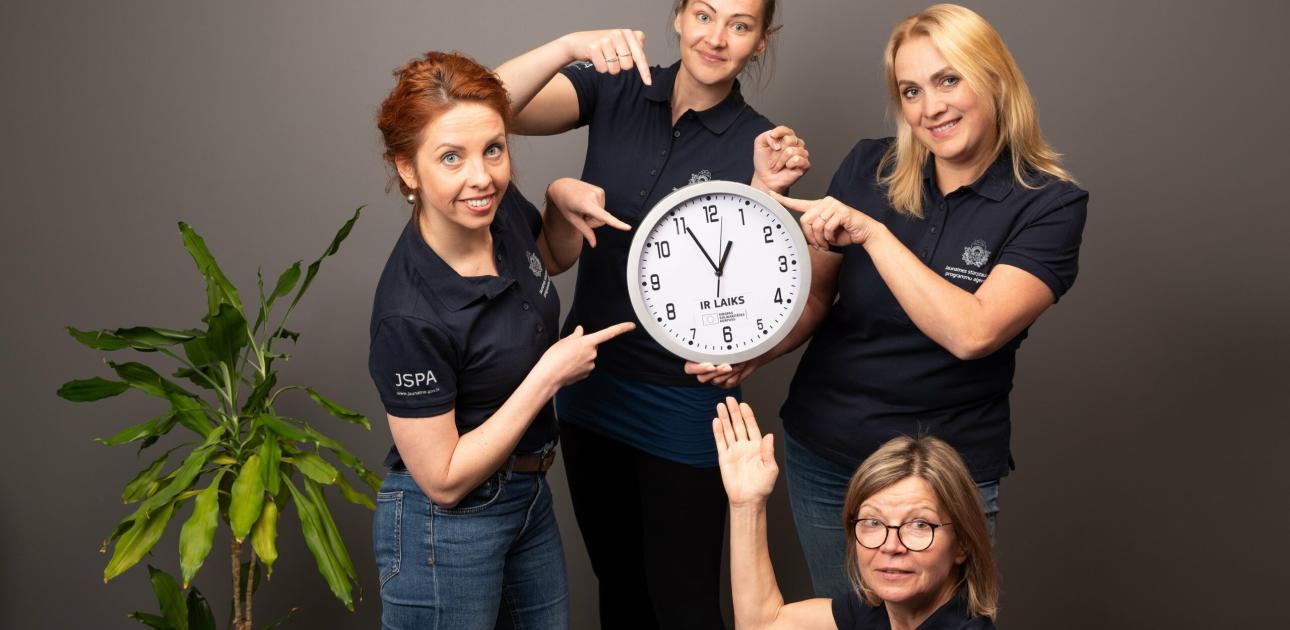 Četras sievietes vienādos krekliņos tur pulksteni un rāda uz to