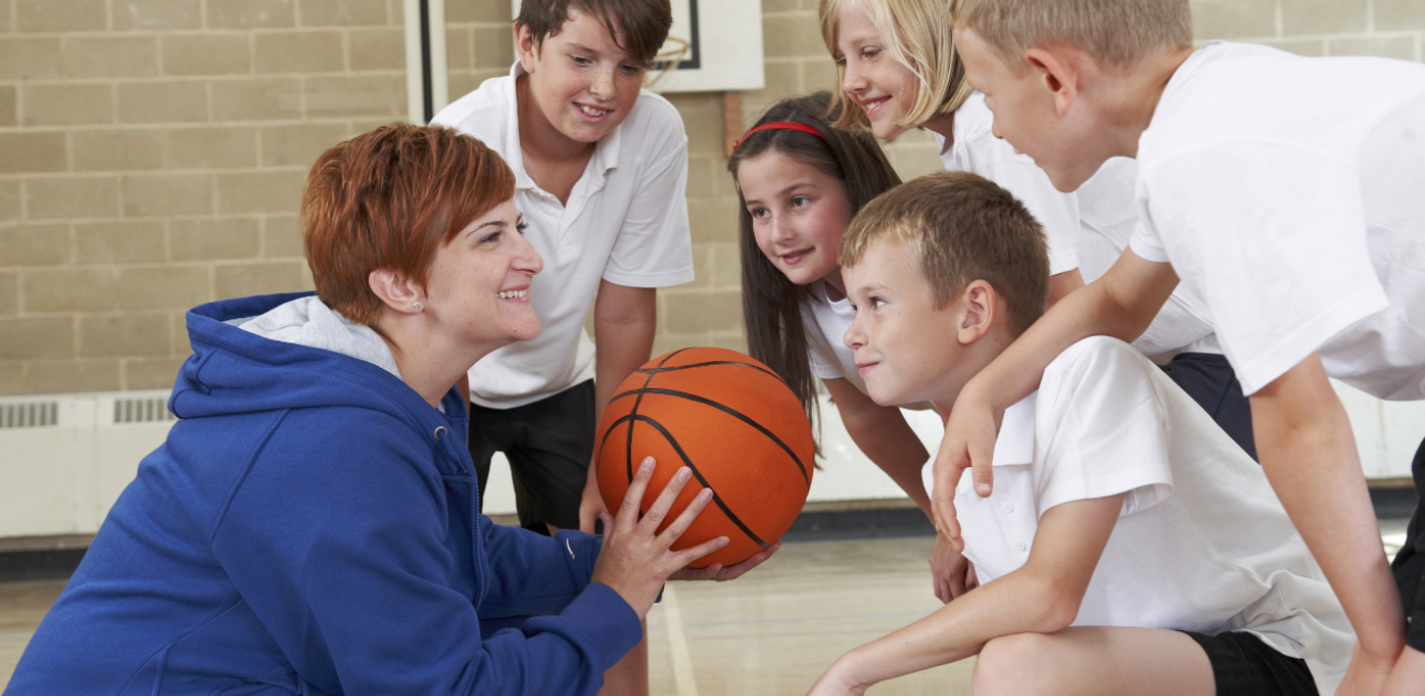Sporta skolotāja ar basketbola bumbu rokās un skolēniem