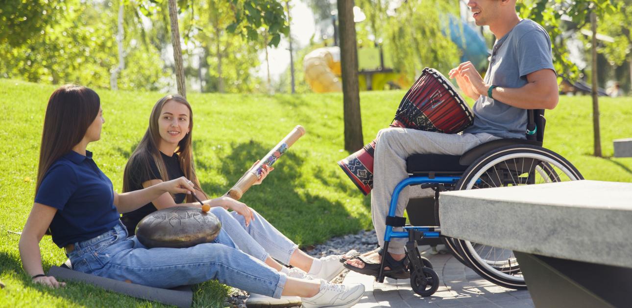 attēlā redzams vīrietis ratiņkrēslā ar mūzikas instrumentu un divas meitenes, kas sēž zālē ar mūzikas instrumentiem