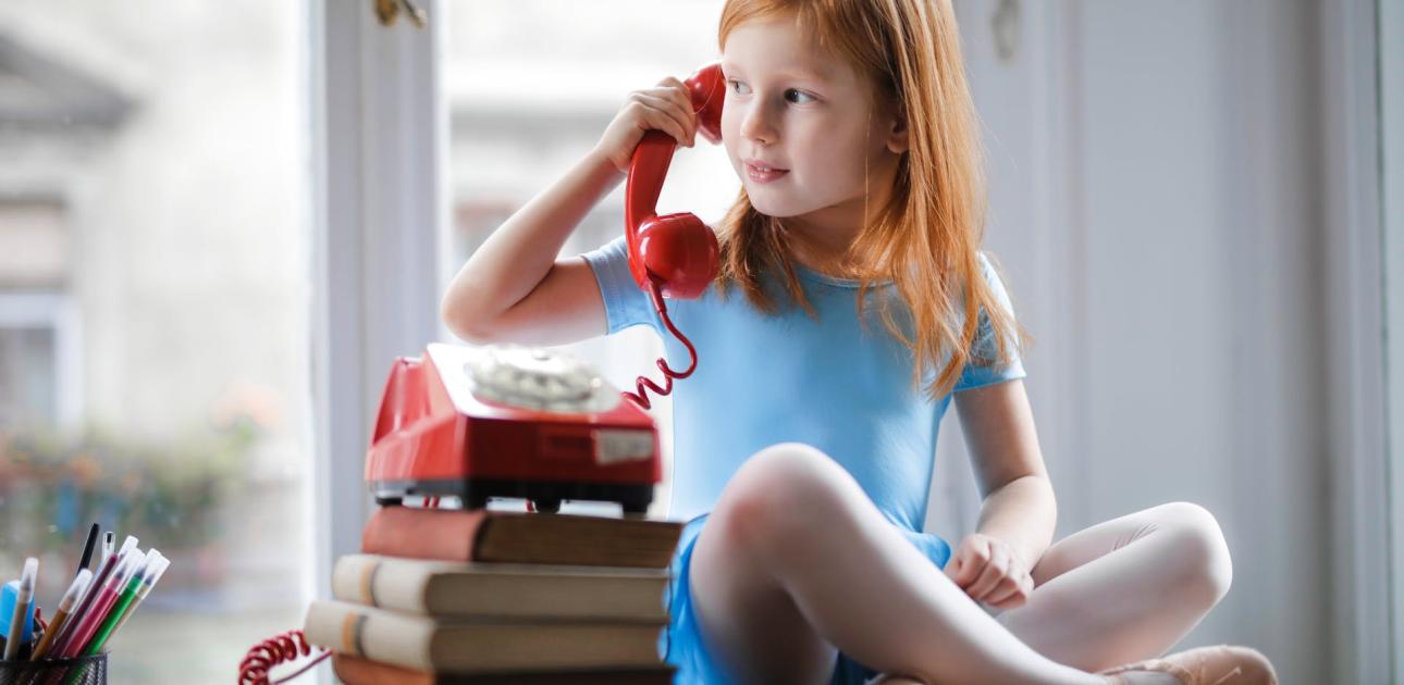 meitene sēž uz galda sakrustojusi kājas un runā pa sarkanu stacionāro telefonu, kas novietots uz grāmatu kaudzes