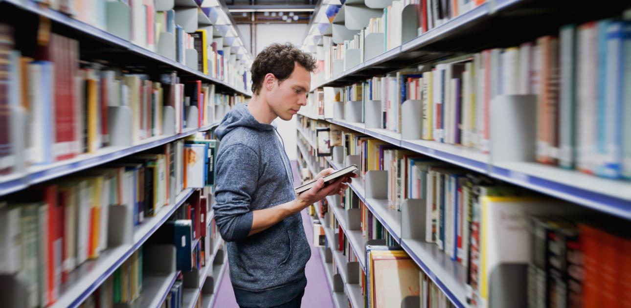 Vīrietis, students ar grāmatu rokās starp diviem grāmatu plauktiem bibliotēkā
