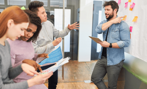 Viens cilvēks stāv pie tāfeles uz kuras ir krāsainas līmlapiņas, trīs cilvēki stāv viņam pretī