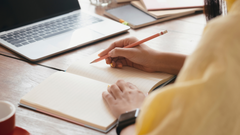 Atvērts piezīmju bloknots, portatīvais dators, sieviete dzeltenā džemperī pieraksta kaut ko 