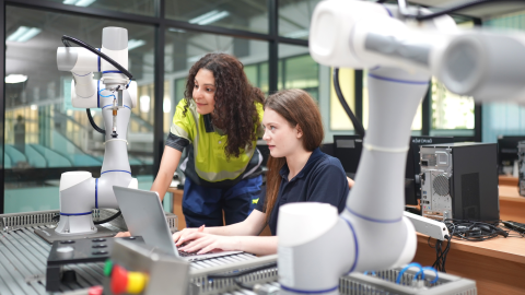 jauna meitene sēž pie datora, viņai blakus kaut ko stāsta pedagogs sieviete, uz galda blakus stāv robototehnika, viņi atrodas mācību darbnīcā