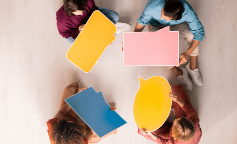 Skats no augšas uz četriem cilvēkiem, kuri sēž uz grīdas un tur rokās krāsainus runas burbuļus