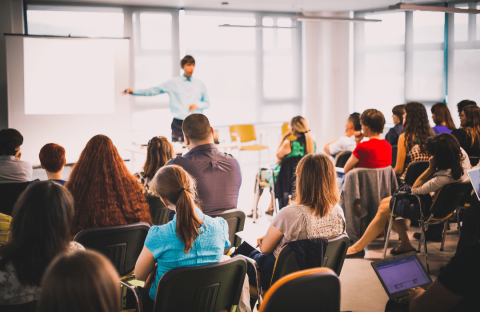 Attēlā redzami cilvēki, kas sēž un klausās runātājā