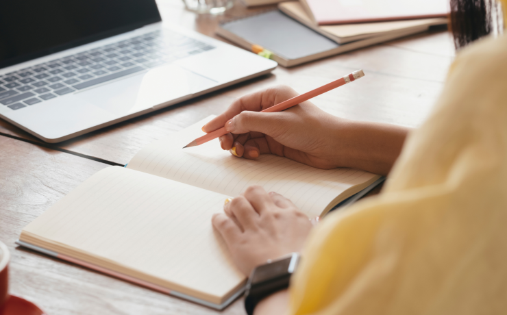 Atvērts piezīmju bloknots, portatīvais dators, sieviete dzeltenā džemperī pieraksta kaut ko 